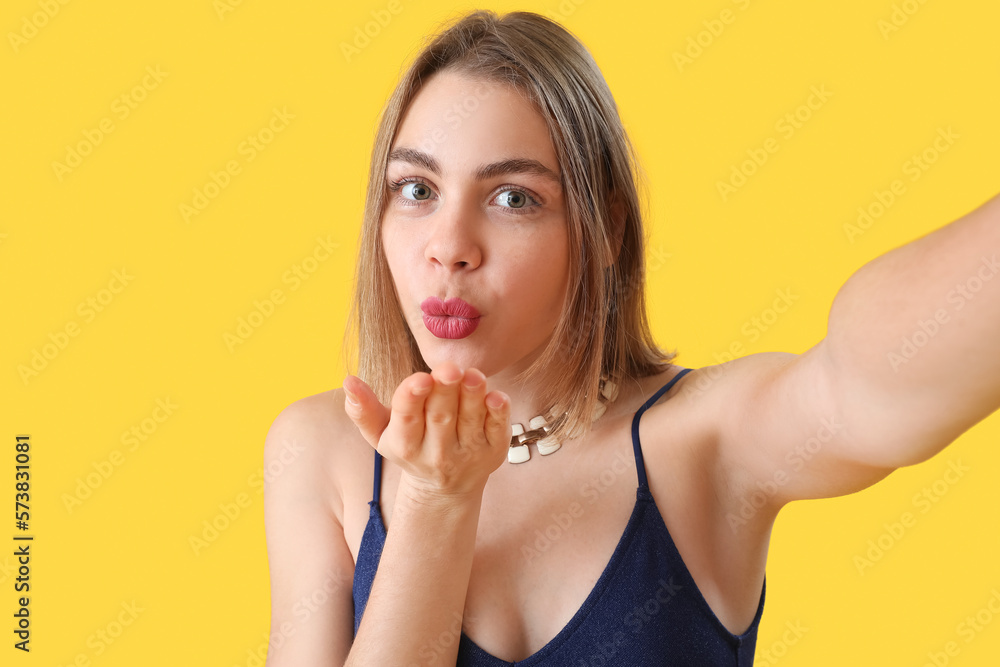 Young woman with necklace blowing kiss on yellow background, closeup