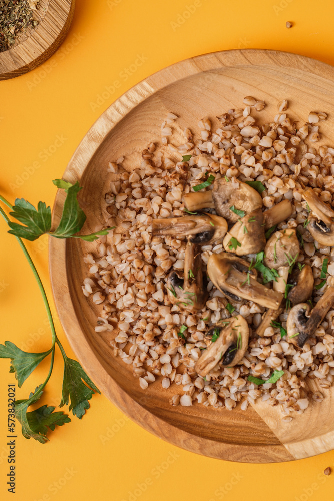 Wooden plate of tasty buckwheat porridge with mushrooms, parsley and dill on yellow background