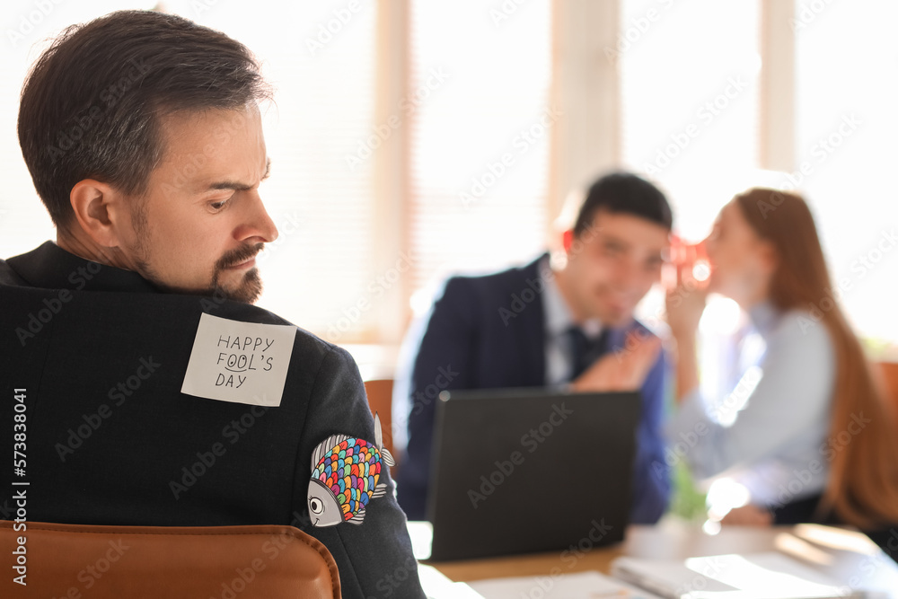 Sticky note and paper fish on back of angry young man in office. April Fools Day celebration