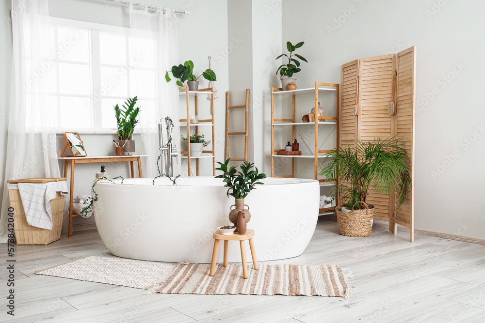 Interior of light bathroom with bathtub, shelving units and houseplants