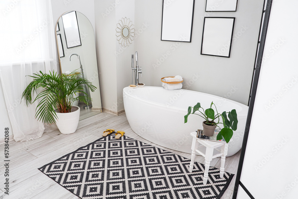 Interior of light bathroom with bathtub, blank frames and houseplants