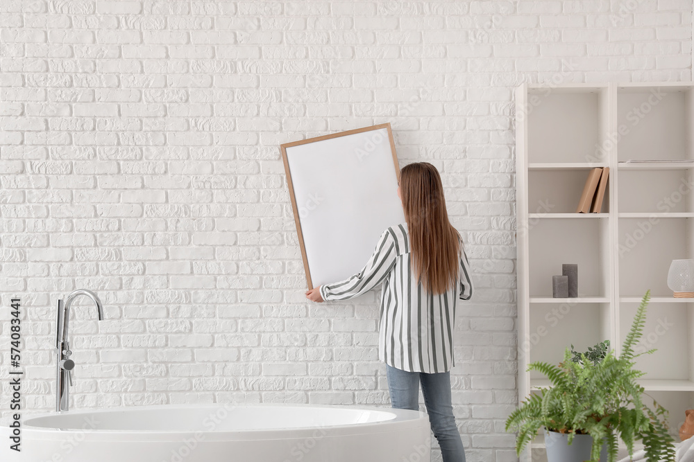 Young woman hanging blank frame on white brick wall in bathroom