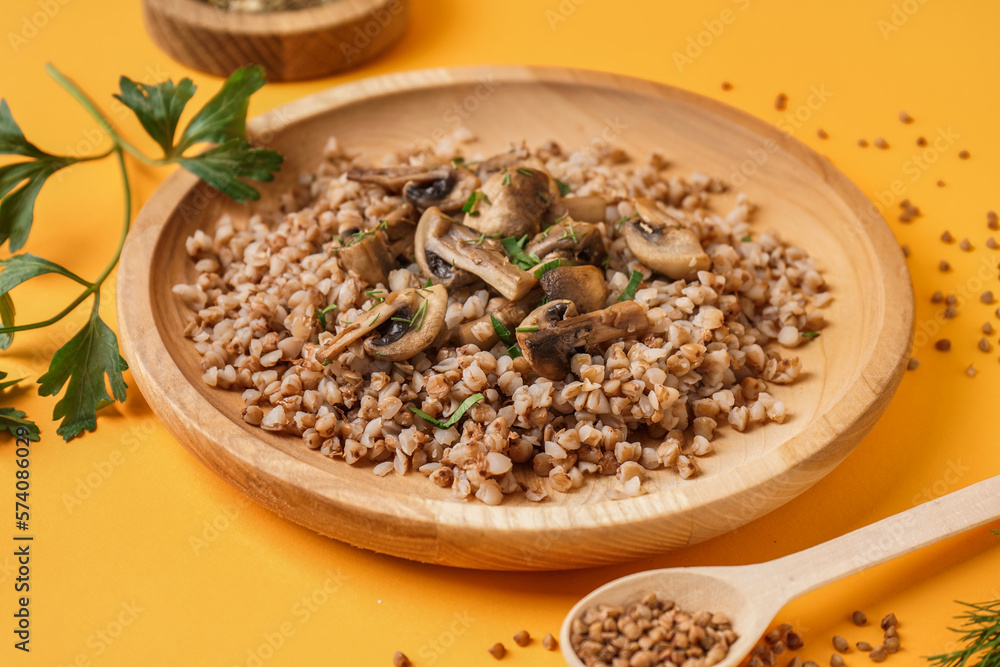 Wooden plate of tasty buckwheat porridge with mushrooms, parsley and dill on yellow background