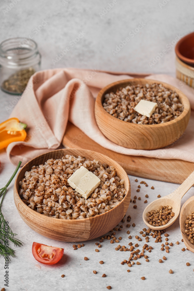 Wooden bowls of tasty buckwheat porridge with butter and vegetables on grey table