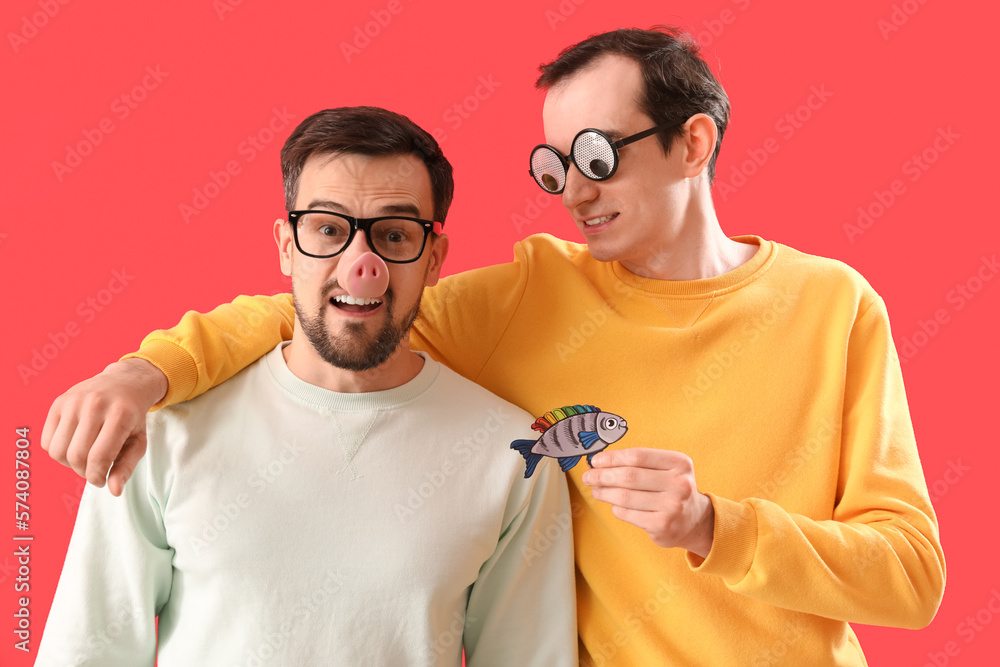 Young men in funny disguise with paper fish on red background. April Fools Day celebration