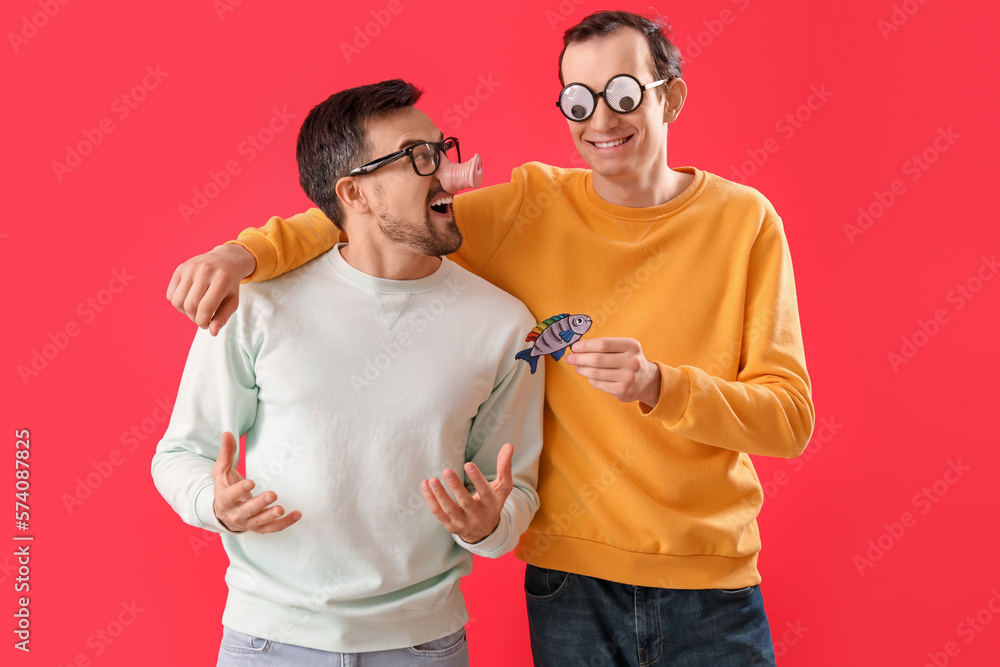 Young men in funny disguise with paper fish on red background. April Fools Day celebration