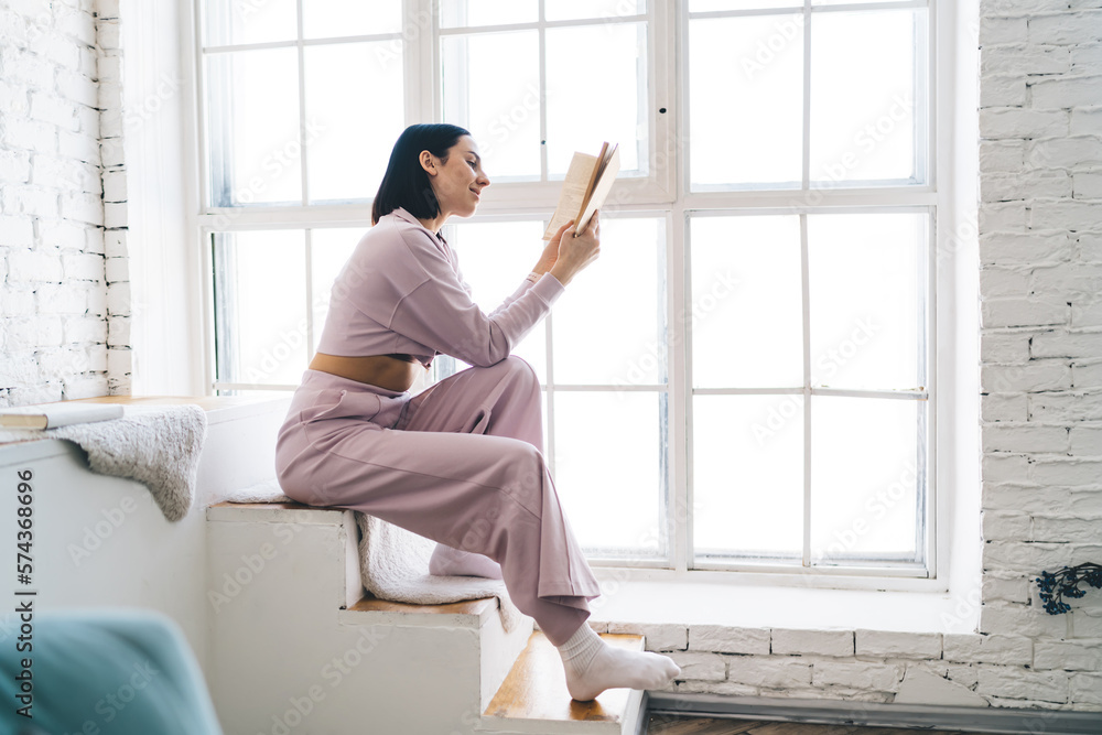 Young woman reading book while sitting near window