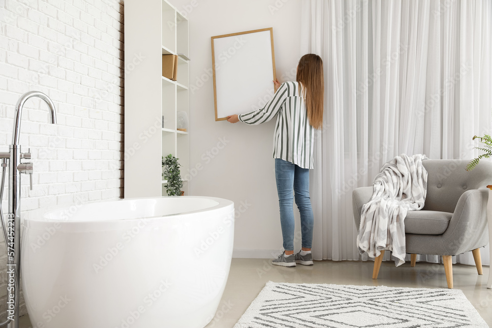 Young woman hanging blank frame on white brick wall in bathroom