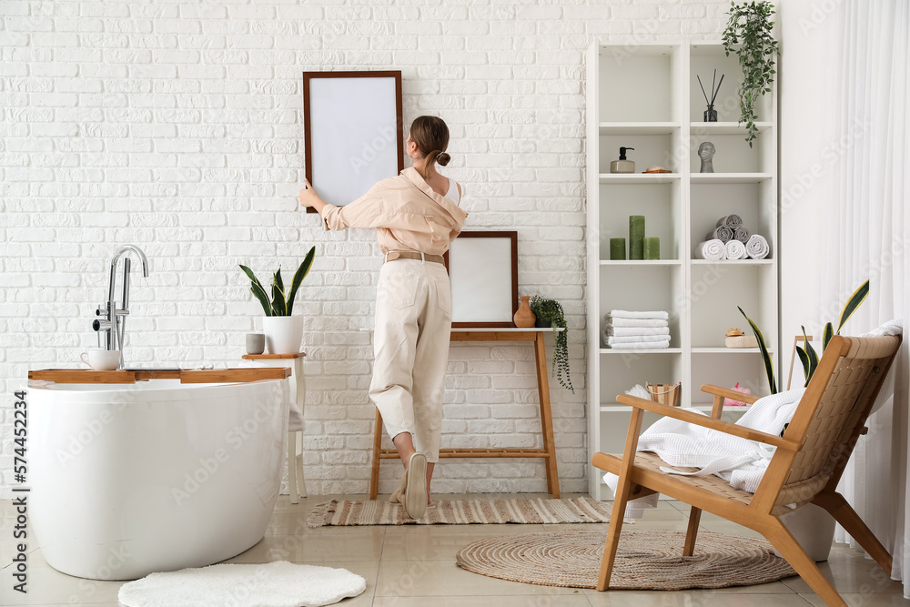 Young woman hanging blank frame on white brick wall in bathroom, back view