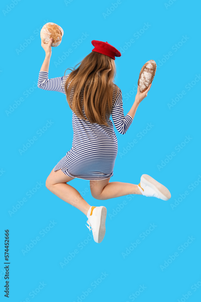 Young woman in beret with loaves of fresh bread jumping on blue background, back view