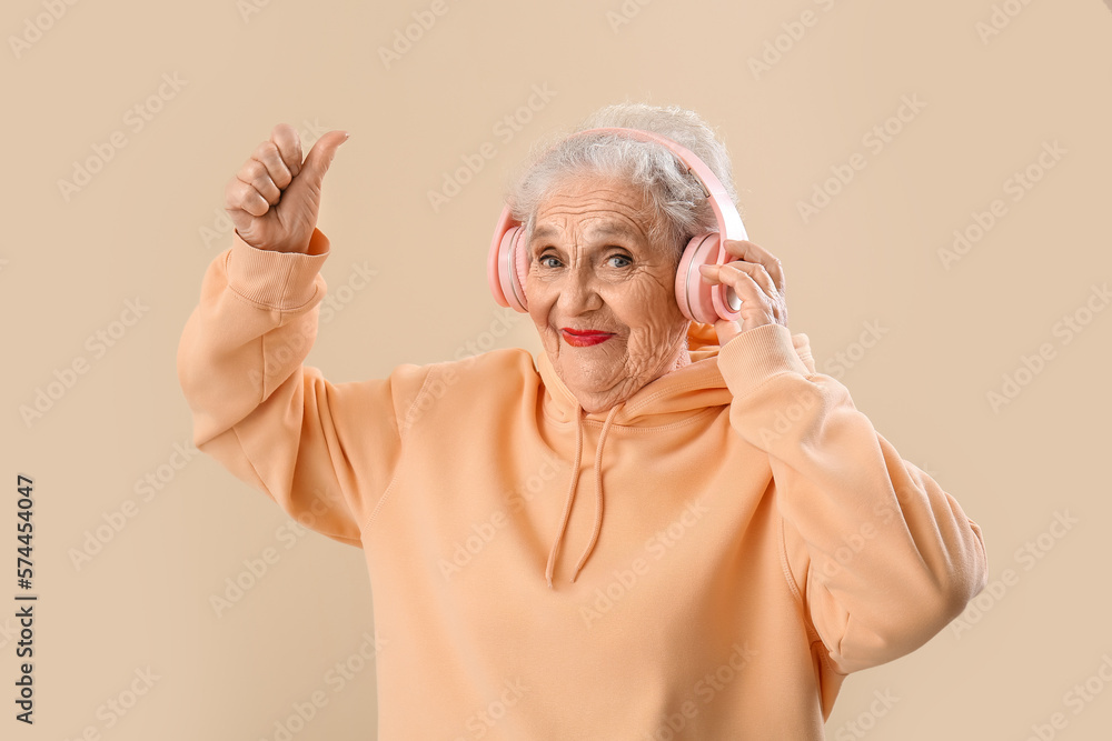 Senior woman in headphones showing thumb-up on beige background