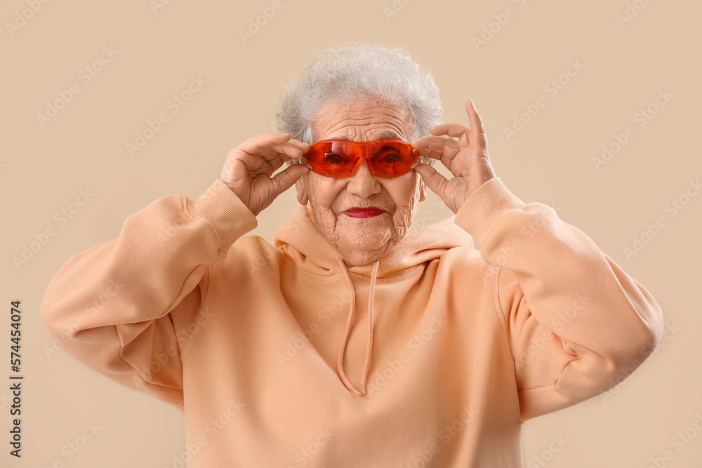 Senior woman in sunglasses and hoodie on beige background