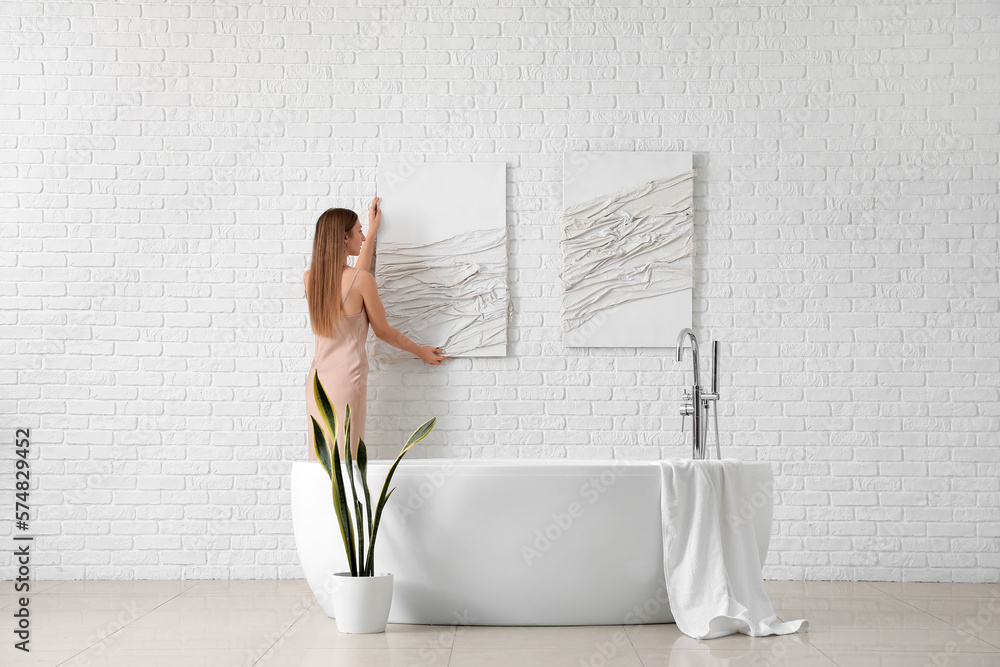 Young woman hanging 3D picture on white brick wall in bathroom