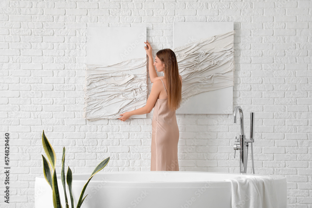 Young woman hanging 3D picture on white brick wall in bathroom