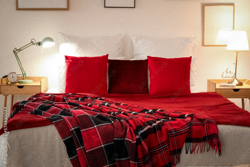 Interior of bedroom with red checkered blankets on bed, blank frames and glowing lamps late in eveni