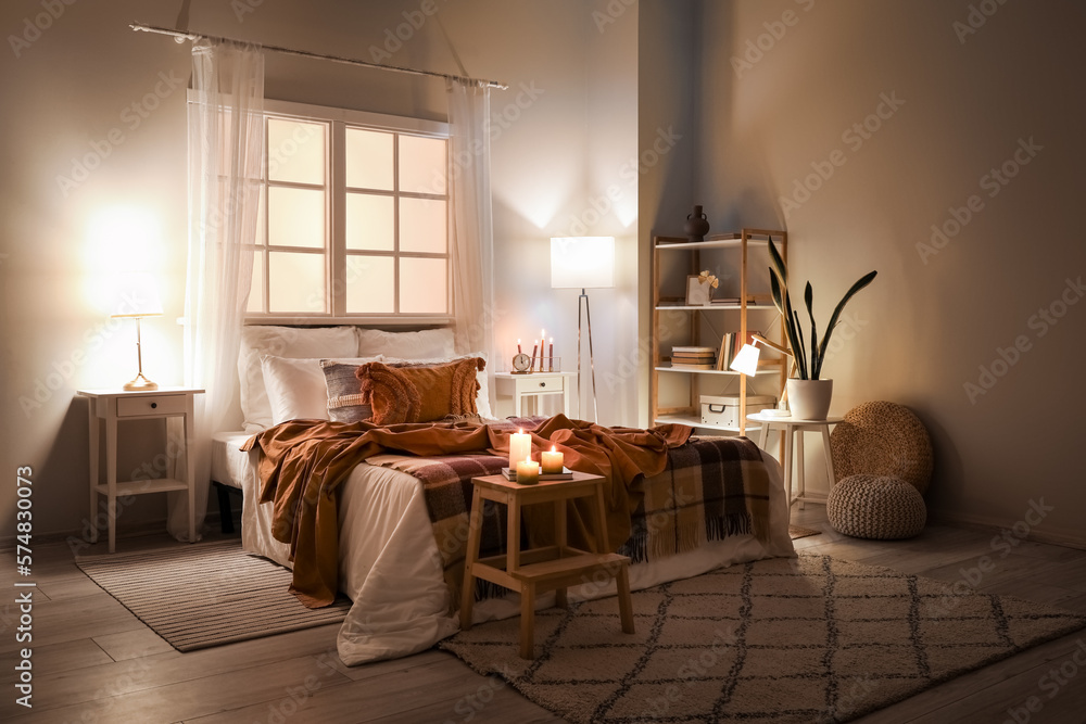 Interior of bedroom with brown checkered blankets on bed, burning candles and glowing lamps late in 