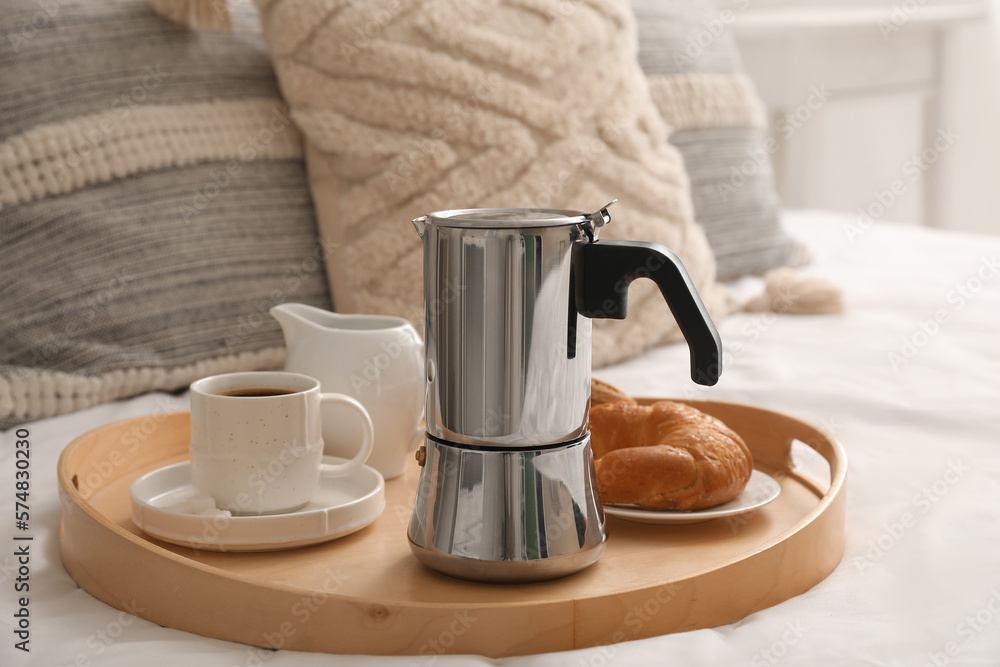 Tray with geyser coffee maker, cup of espresso and delicious croissant on bed