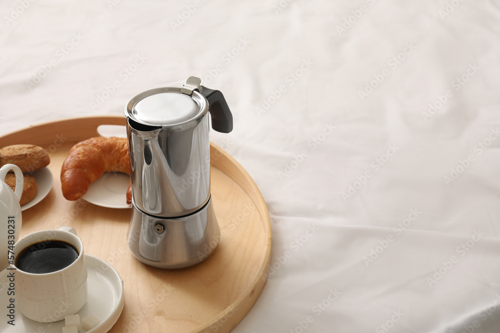 Tray with geyser coffee maker, cup of espresso and delicious croissant on bed