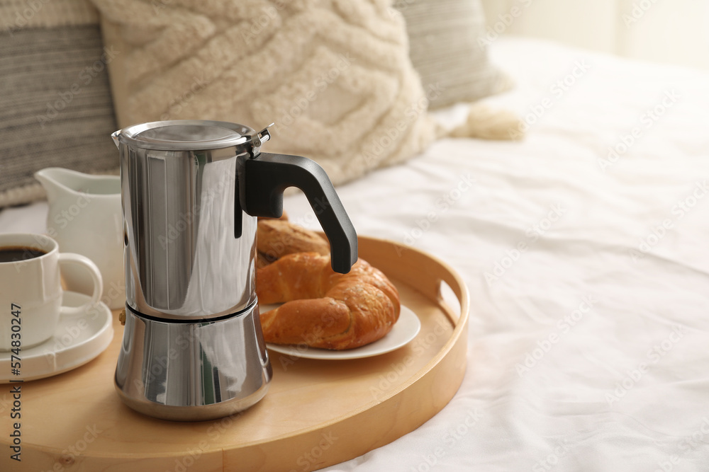 Tray with geyser coffee maker, cup of espresso and delicious croissant on bed