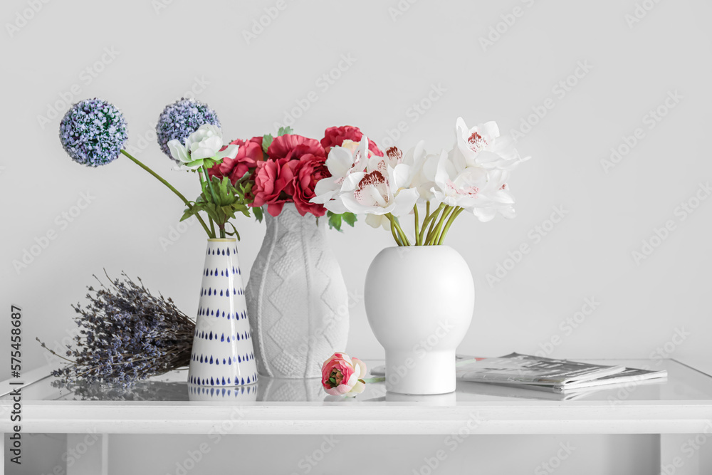 Vases with beautiful autumn flowers and magazines on table near light wall