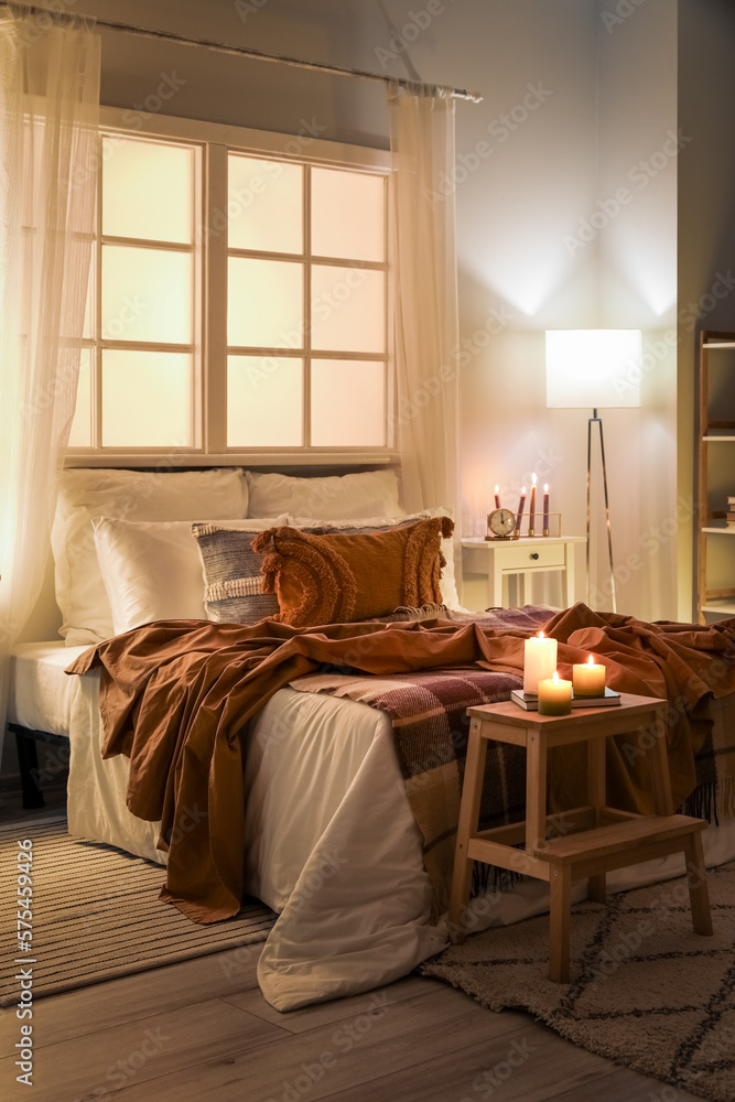 Interior of bedroom with brown checkered blankets on bed, burning candles and glowing lamp late in e