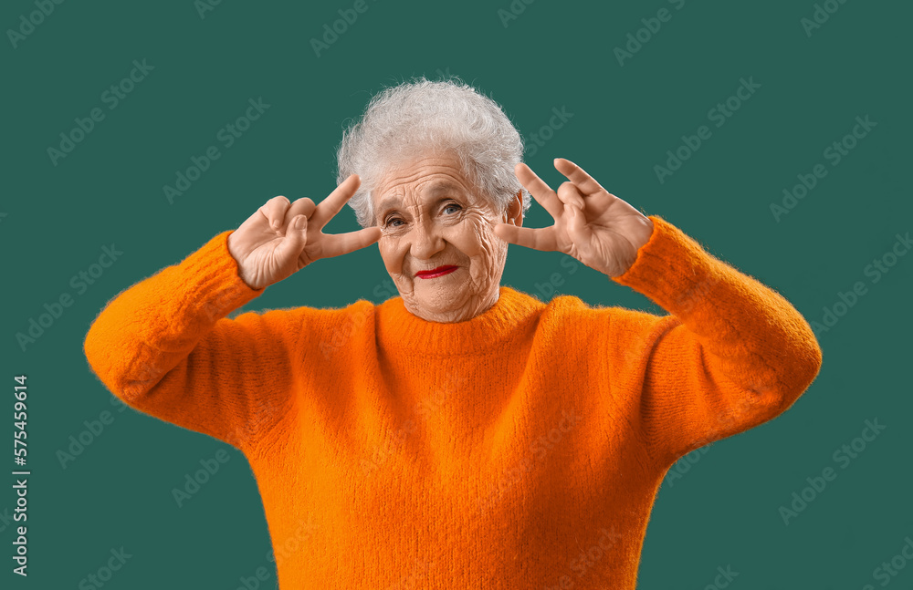 Senior woman in orange sweater showing victory gesture on green background