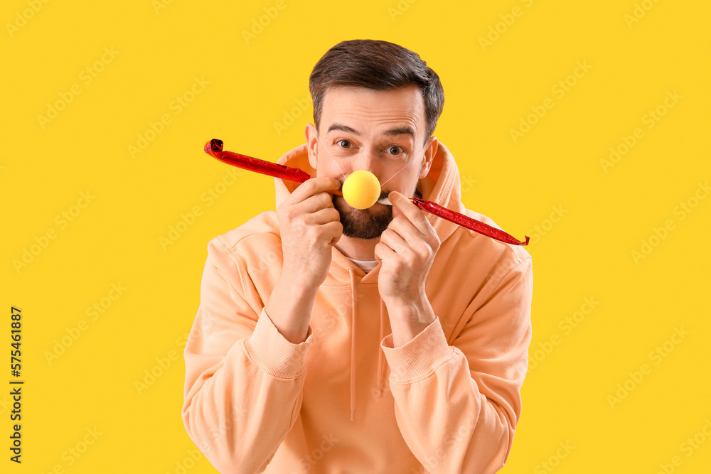 Funny young man with clown nose and party blowers on yellow background. April Fools Day celebration