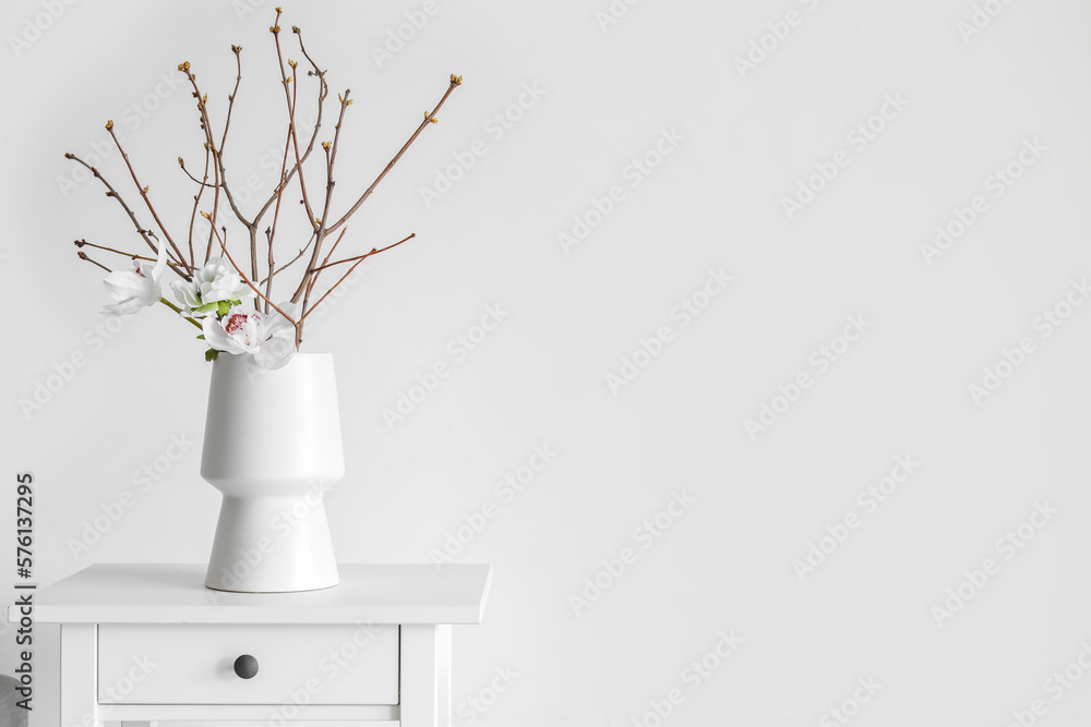 Vase with tree branches and flowers on table near light wall