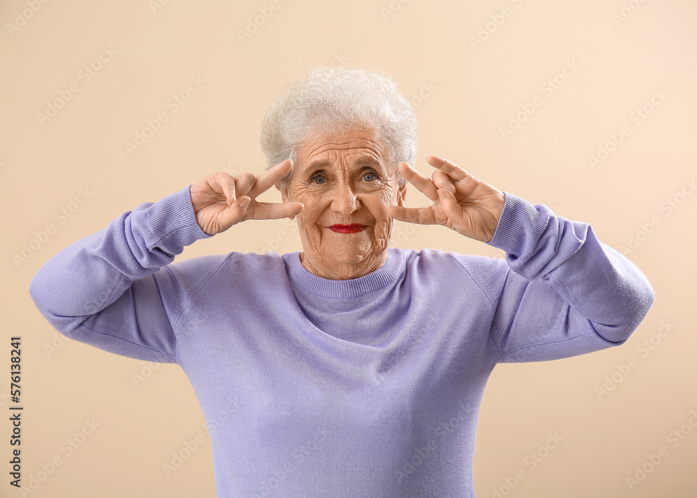 Senior woman in lilac sweater showing victory gesture on beige background