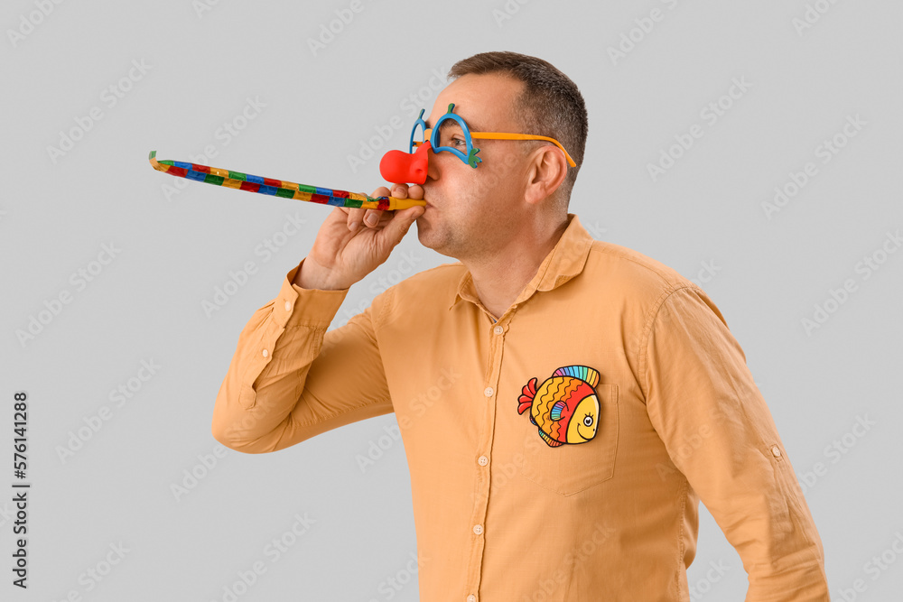 Mature man in funny disguise with party blower on grey background. April Fools Day celebration