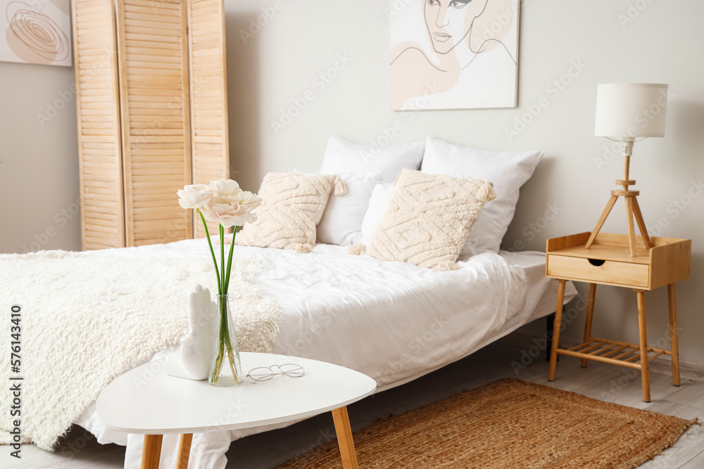 Vase with ranunculus flowers and eyeglasses on table in interior of bedroom