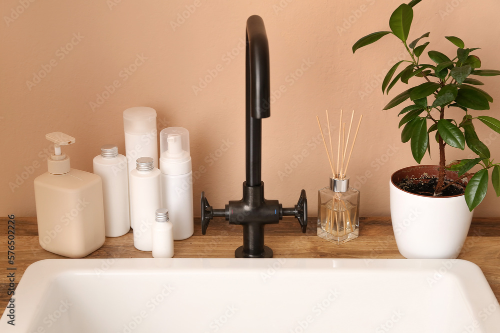 Table with sink, cosmetic products, reed diffuser and houseplant near beige wall