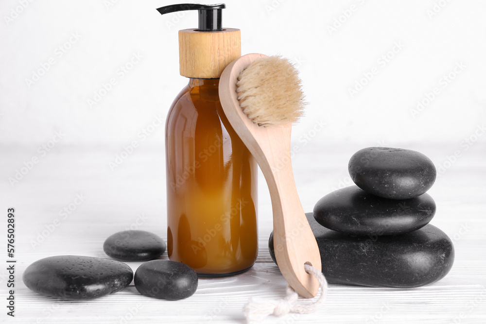 Massage brush, bottle of cosmetic product and spa stones on light wooden table