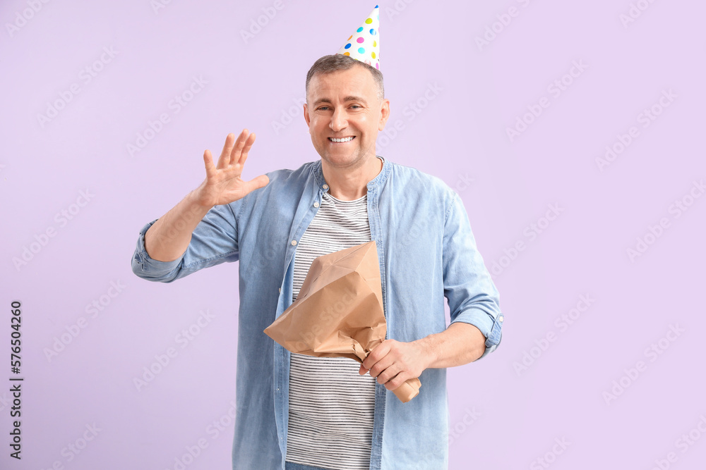 Mature man in party hat with bag on lilac background. April Fools Day celebration