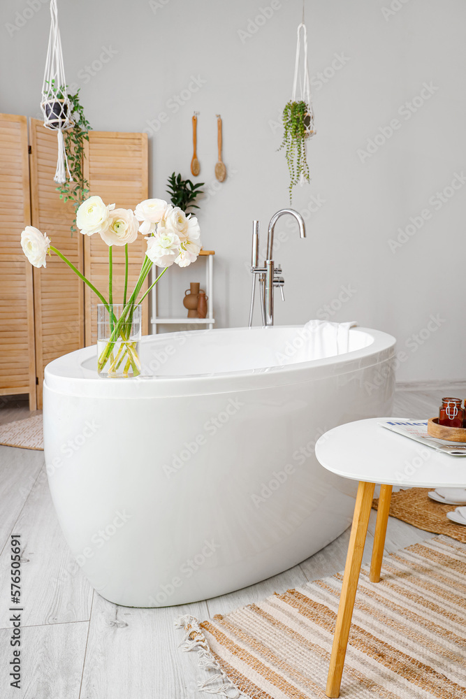 Vase with ranunculus flowers on bathtub in interior of light bathroom