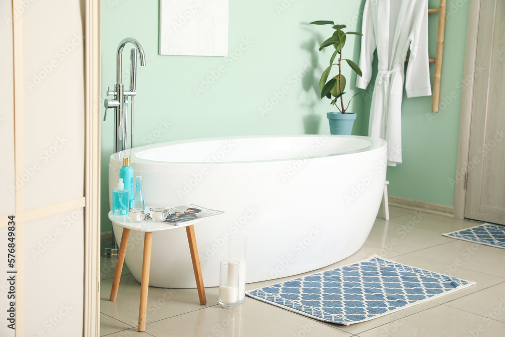 Interior of bathroom with modern bathtub and bath accessories near green wall