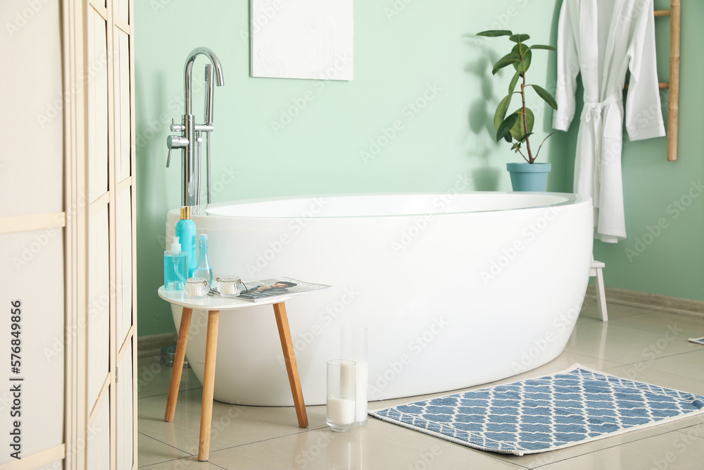 Interior of bathroom with modern bathtub and bath accessories near green wall