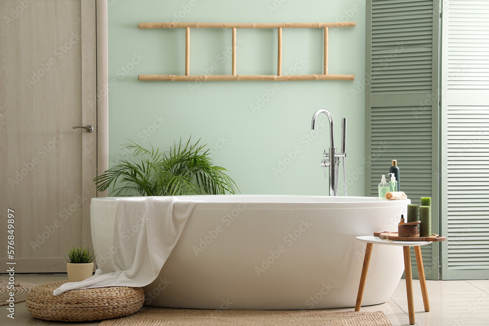 Interior of bathroom with modern bathtub, bath accessories and houseplants near green wall