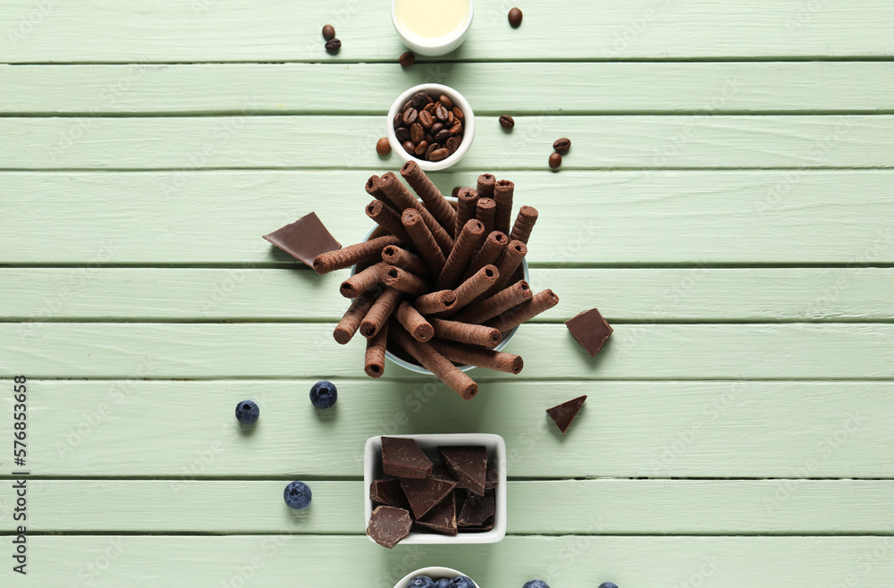 Delicious wafer rolls, chocolate pieces, coffee beans and blueberries on green wooden background