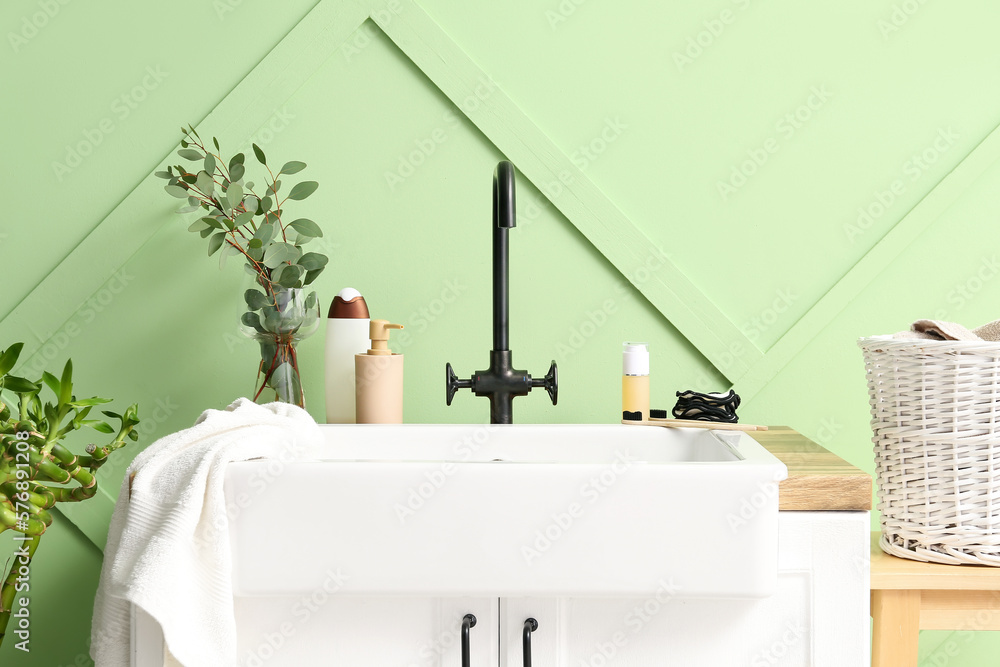 Table with ceramic sink, bath accessories and eucalyptus in vase near green wall