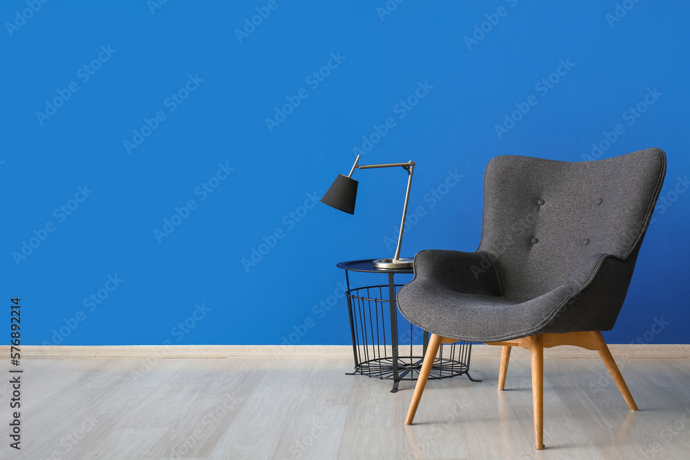 Stylish grey armchair and lamp on end table near blue wall