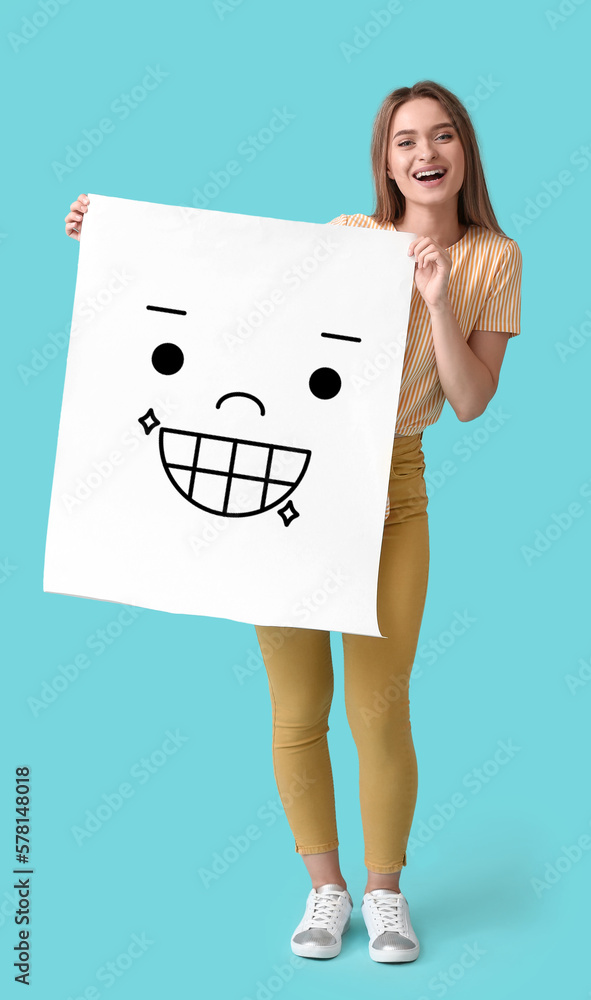 Happy young woman holding paper with smiling emoticon on light blue background