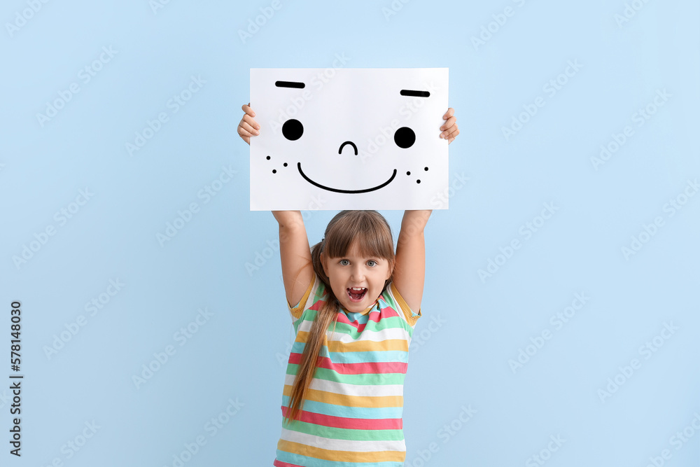 Happy little girl holding paper with smiling emoticon on light blue background