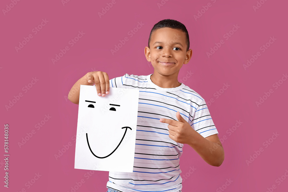 Little African-American boy holding paper with happy emoticon on pink background