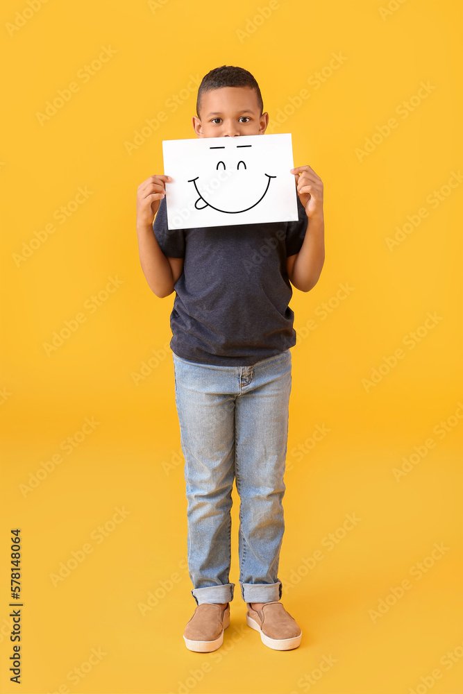 Little African-American boy holding paper with happy emoticon on yellow background