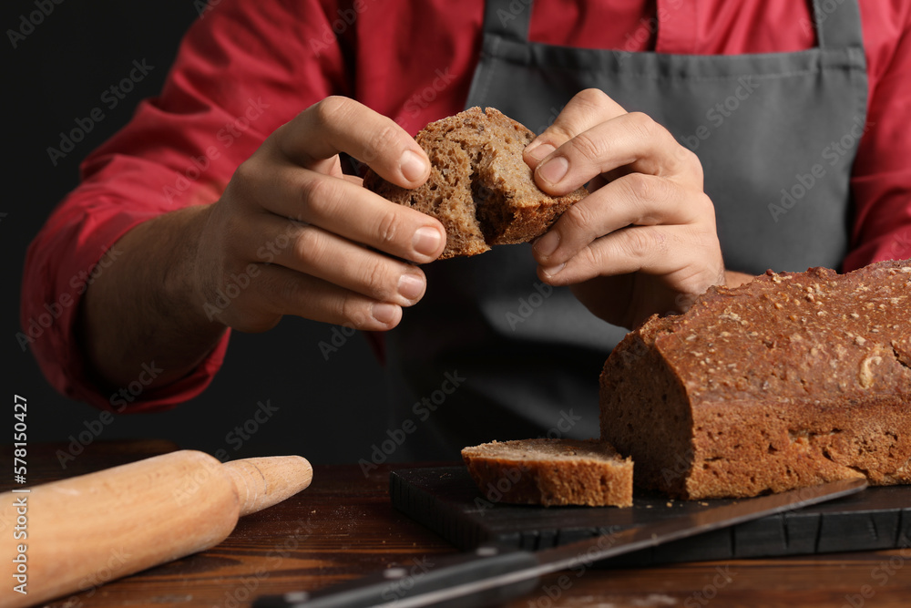 Male baker with slice of rye bread at table on dark background, closeup