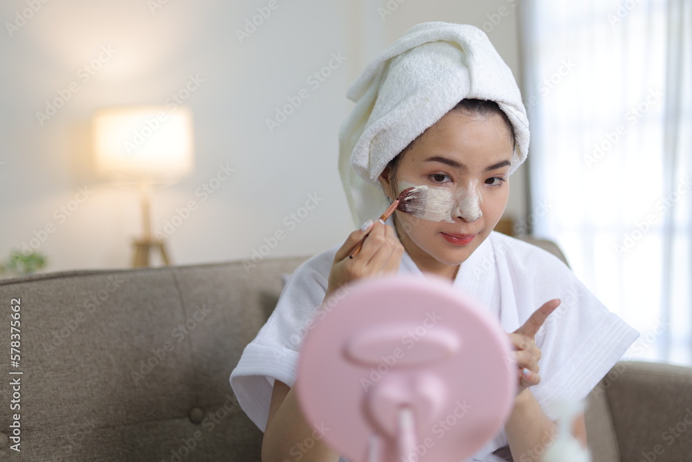 Happy young asian woman wrapped in towel applying skin cream on face. smile and look in the mirror, 