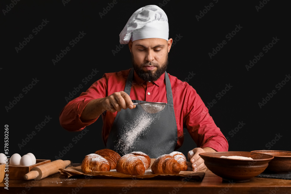 Male baker sprinkling croissants with sugar powder at table on dark background