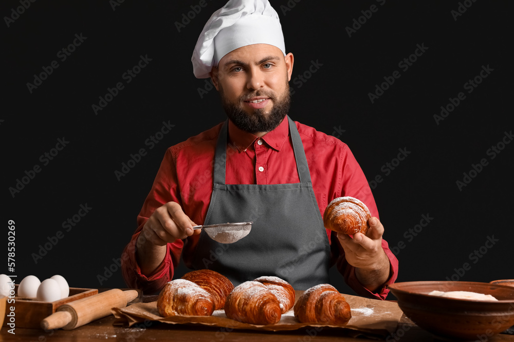 Male baker sprinkling croissants with sugar powder at table on dark background