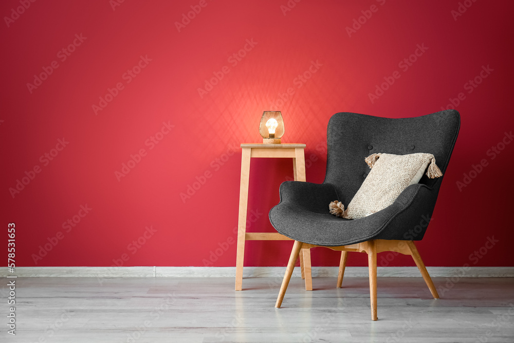 Stylish grey armchair, cushion and end table with glowing lamp near red wall